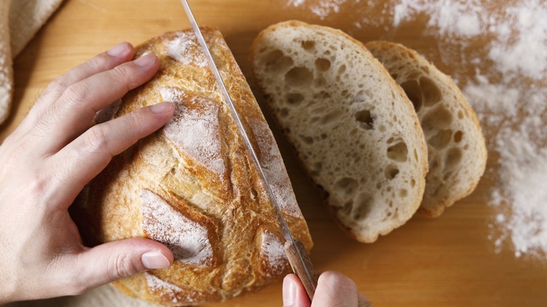 Hands using a knife to slice sourdough bread on floured surface