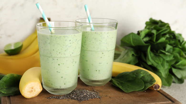 Two green smoothies in glass cups with paper straws on a wooden cutting board with chia seeds, spinach, banana, and lime