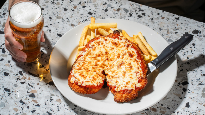 Plate of chicken parmigiana and French fries next to a hand holding a beer