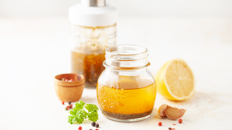 A glass jar of Italian salad dressing styled with fresh ingredients on a white background