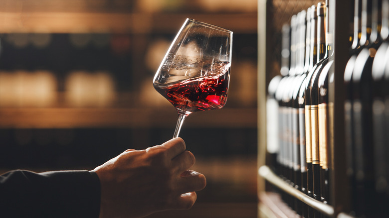 A man swirls red wine in a stem glass next to shelves of bottles