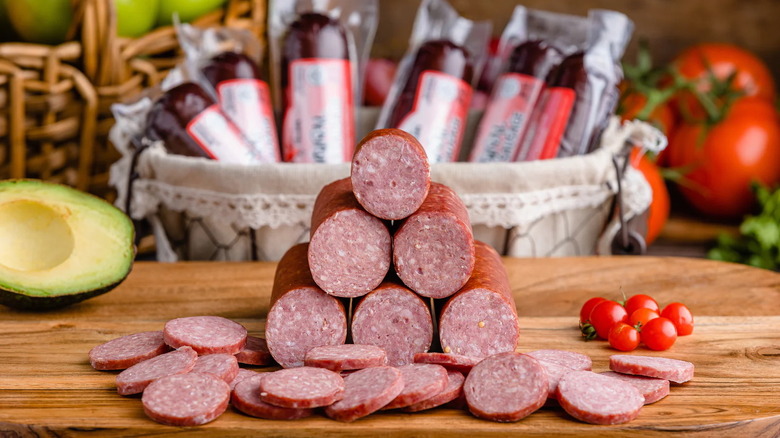 Echo Valley Meats sliced sausages placed on top of wooden chopping board while a sliced avocado, tomatoes, and other packed ingredients are displayed at the back.