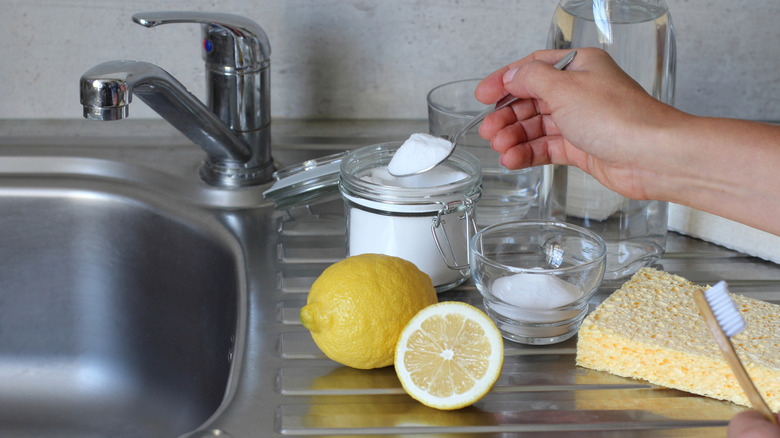 A had scooping salt with a spoon, surrounded by lemon, water, a sponge, and a toothbrush, next to a sink