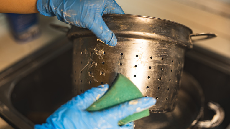 Gloved hands cleaning a stainless steel strainer with a sponge