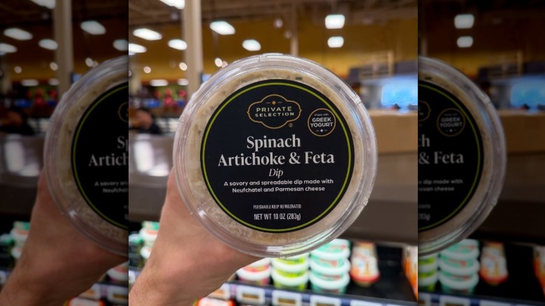 A hand holding a tub of Kroger Private Selection Spinach Artichoke & Feta Dip in the store.