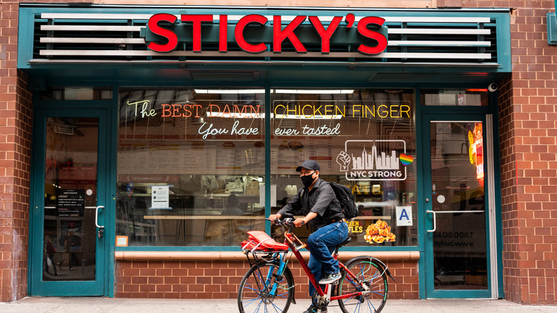 The exterior of a Sticky's Chicken Joint location in New York City as a man wearing a mask rides a bicycle in front of it.