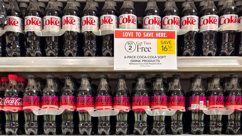 Plastic bottles of Diet Coke and Coca-Cola Cherry Zero Sugar on a supermarket shelf, with a promotion sign offering "Buy 2, Get 2 Free" on six-packs.