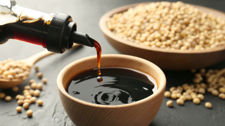 Bottle pouring soy sauce into wooden bowl with dish of soy beans in the background