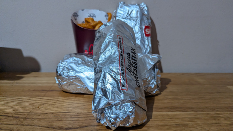 Three foil-wrapped Arby's cheesesteaks with fries in the background