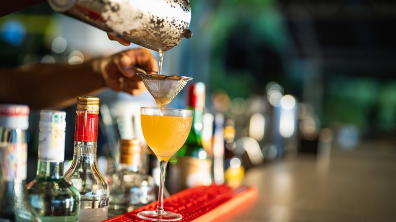 Bartender double strains a cocktail into a glass at a bar.