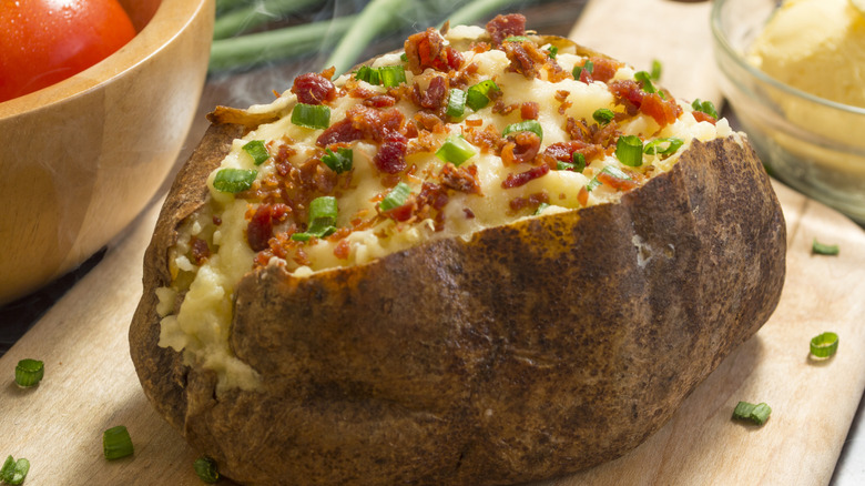 A big baked potato split open with chives and bacon bits on top sitting on a wood cutting board.