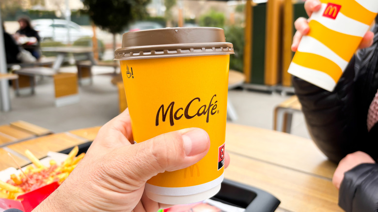 A hand holds up a McCafé coffee cup while another person drinks from a regular McDonald's cup in the background