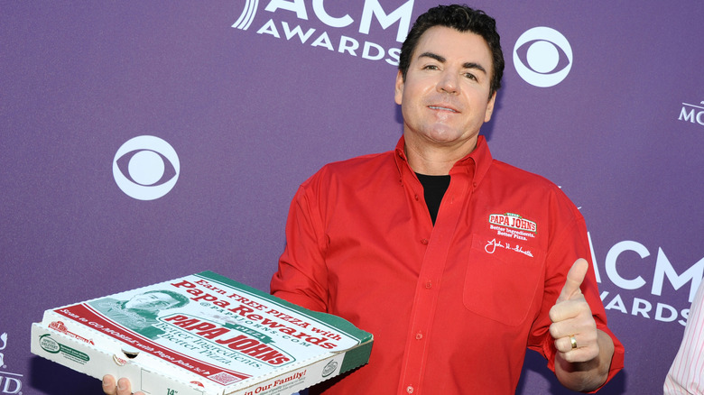 John Schnatter, in a red Papa Johns button down, holds a Papa Johns pizza box in front of a purple backdrop with white text.