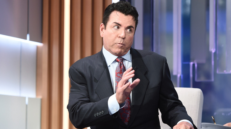 John Schnatter, with dark hair in a dark suit with a red tie, talks animatedly on the sent of a news show.
