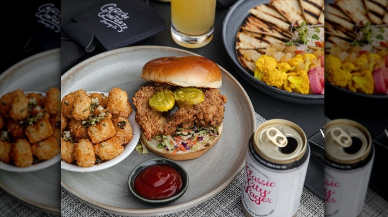 a plate with a chicken sandwich with pickles and slaw with a side of tater tots and ketchup. Behind is another plate with a quesadilla and broccoli on the side