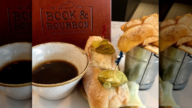 Short Rib Dip sandwich with cheese, a cup of au jus, and a side of chips. A red menu that reads "book & bourbon southern kitchen" sits in the back