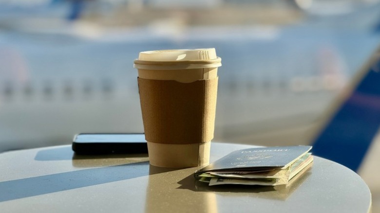 a coffee cup, phone, and passport sit on a table in front of a window where a plane is seen parked on the runway