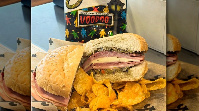 a muffuletta sandwich with meat, cheese, and olive salad on a sesame bun, with a bag of New Orleans Voodoo kettle chips
