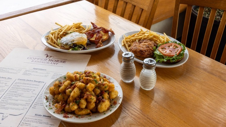 3 plates with burgers, eggs and bacon, fries, and tater tots on a table next to a menu that reads "Copper Horse Restaurant"