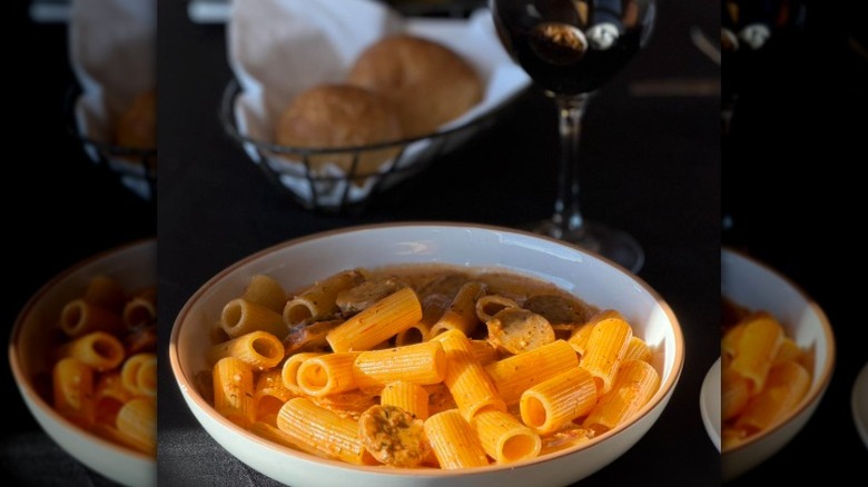 a bowl of rigatoni alla vodka with sausage in front of a bread basket and glass of wine
