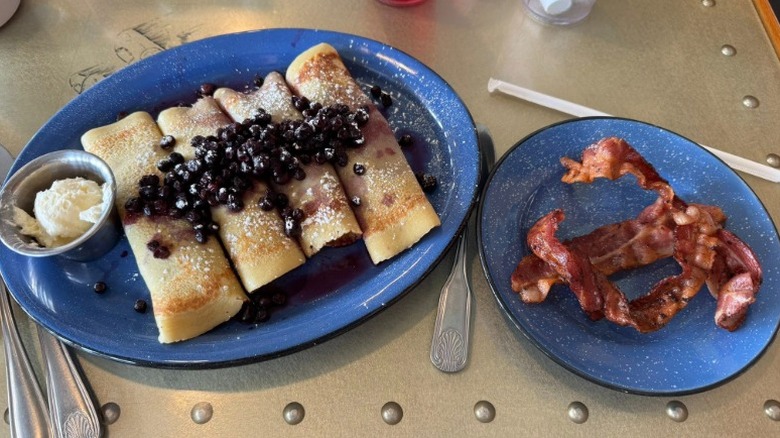 a plate with four crepes with powdered sugar, blueberries, and a side of cream, and a plate with bacon on a table