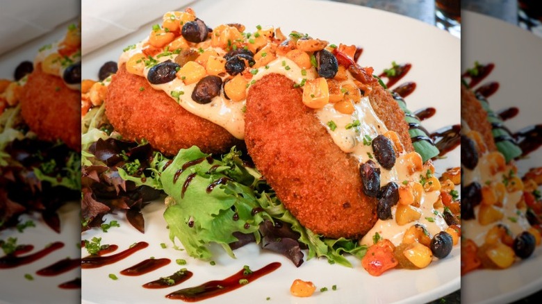 two round fish cakes with a white sauce, corn and beans, and lettuce on a plate