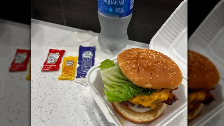 hamburger with cheese and lettuce in a takeout container with sauce packets and a water bottle