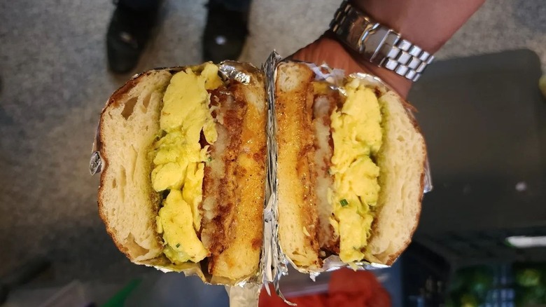 A brimming breakfast sandwich with egg and hash brown being held by a man with a watch