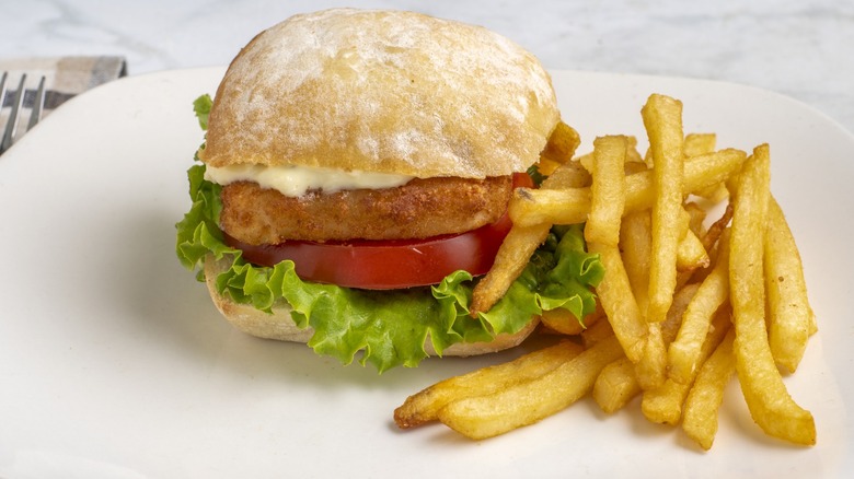 Haddock fillet with lettuce , tomato, and tarter sauce on a ciabatta bun served with french fries on a white plate