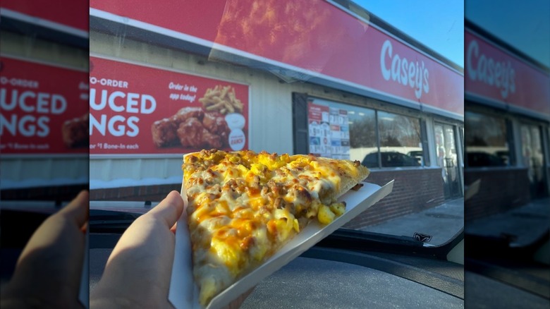 A hand holds up a piece of Casey's breakfast pizza in a car outside the store