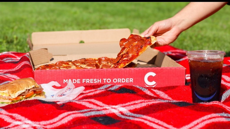A hand reaches for a Crosby's pizza that is on a picnic blanket with soda and a sandwich
