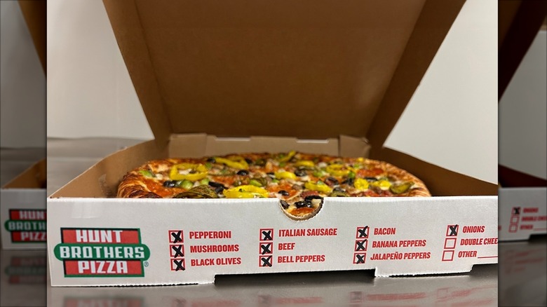 A Hunt Brothers Pizza sits in a box with most of the toppings checked off