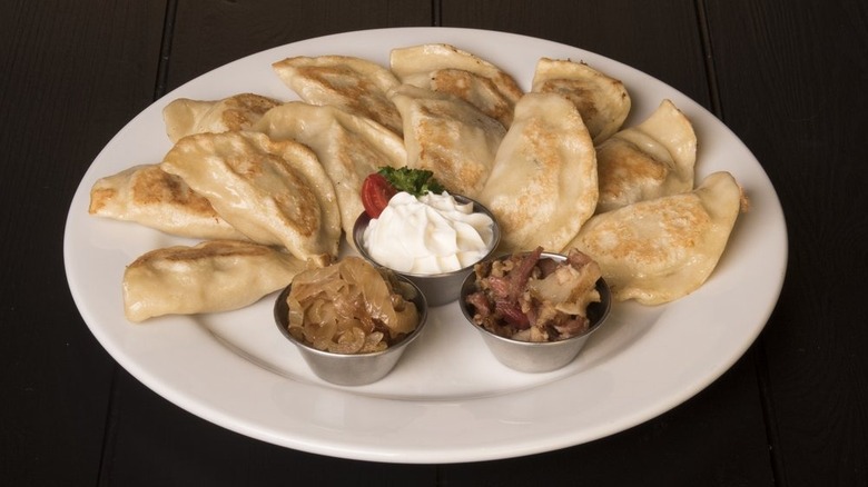 12 pan-fried pierogi on a white plate set on a dark surface with a metal ramekin of sour cream, half a grape tomato, and parsley in the center and metal ramekins of caramelized onions