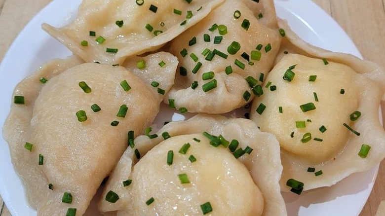 Five boiled pierogi on a white plate that is set on a light wood surface topped with chopped chives