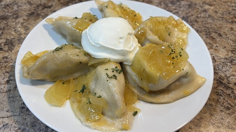 Six pierogi on arranged in a pinwheel shape on a white plate with a dollop of sour cream in the middle and topped with herbs and caramelized onions