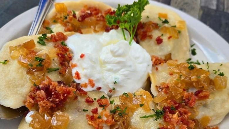 Six pierogi arranged in a circle on a white plate with a dollop of sour cream and parsley spring in the middle, garnished with bacon, caramelized onions, and parsley