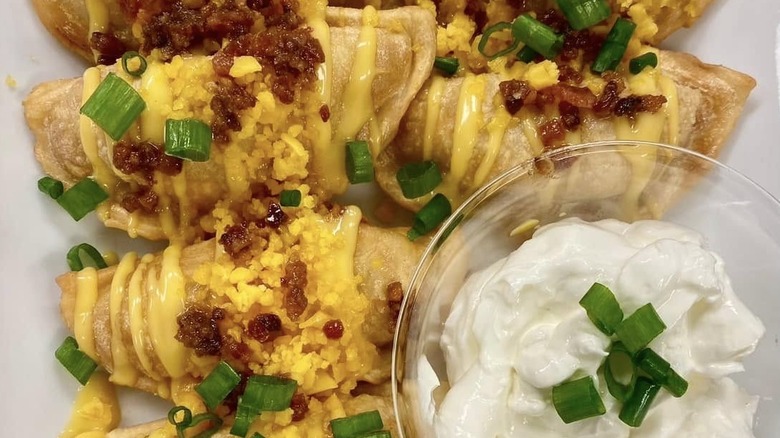 Close-up of pan-fried pierogi on a white plate topped with bacon and chopped green onions and drizzled with a yellow sauce accompanied by a glass ramekin filled with sour cream