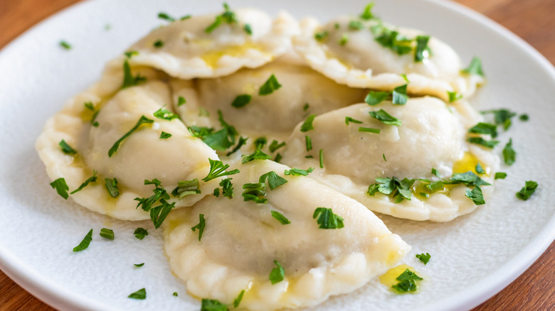 Six boiled pierogi, drizzled with butter and sprinkled with parsley, on a white plate set on a medium-tone wood surface