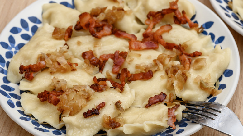 A dozen boiled pierogi topped with chopped bacon and caramelized onions on a white European-style plate with a blue graphic flower border