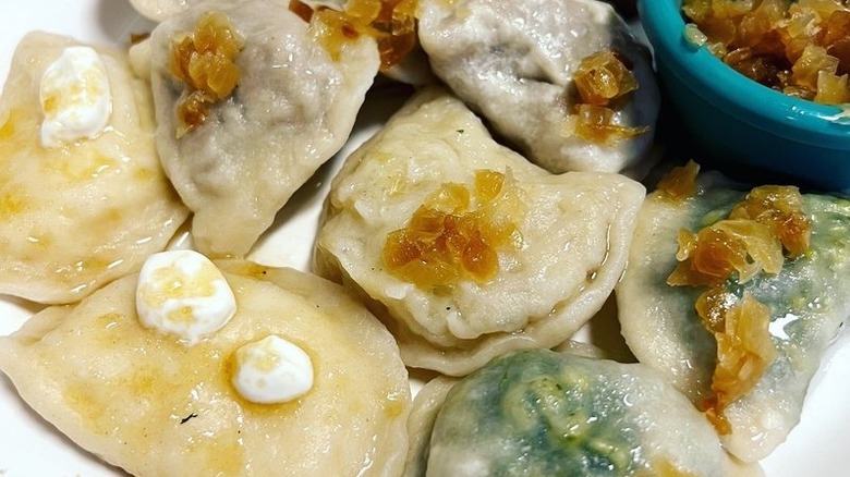 Assorted pierogis on a white plate topped with sour cream and caramelized onions and a blue ramekin of caramelized onions on the side
