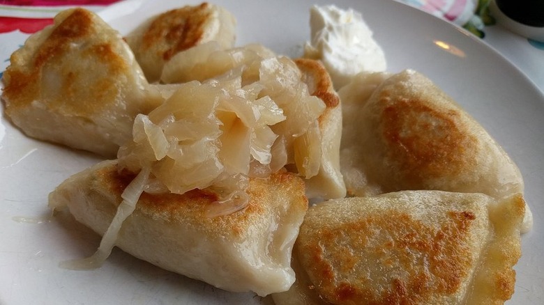 Six pan-fried pierogi on a white place topped with caramelized onions with a dollop of sour cream