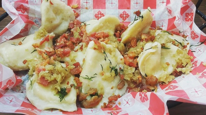 Nine pierogi in a basket lined with red and white checkerboard paper topped with caramelized onions, bacon, and herbs