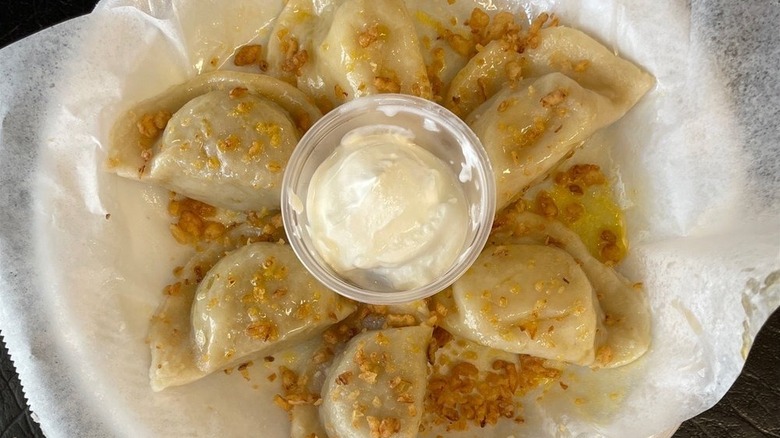 Six pierogi arranged in a fan shap on a white wax paper-lined styrofoam plate set on a black surface with a clear plastic ramekin of sour cream in the center and topped with butter and crispy bits of onion
