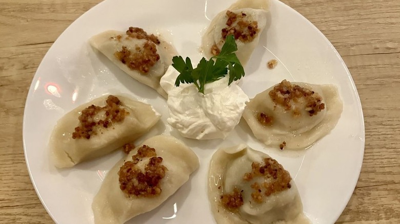 Six boiled pierogi arranged in a fan shape on a white plate on a light wood surface with dollop of sour cream and a parsley sprig in the center and topped with bacon
