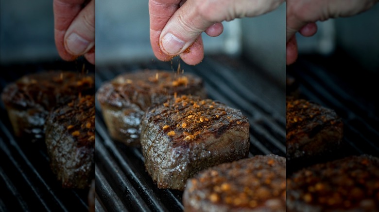 close-up of a chef's hand sprinkling seasoning over steaks on a grill
