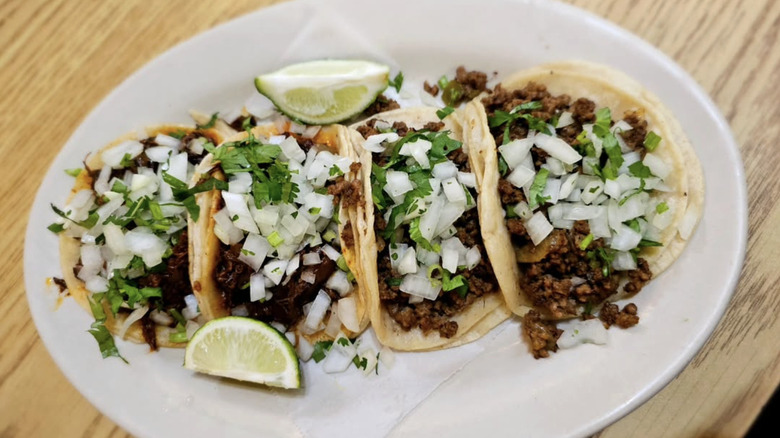 Four tacos with onion, cilantro and lime wedges on a plate