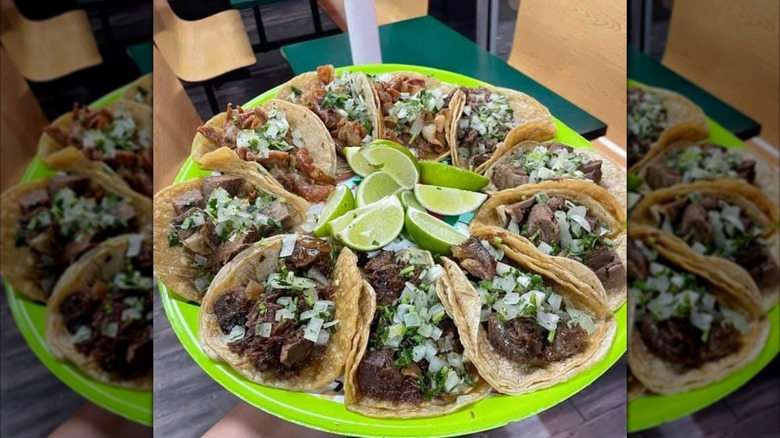A big green paper plate full of different tacos with toppings and lime wedges