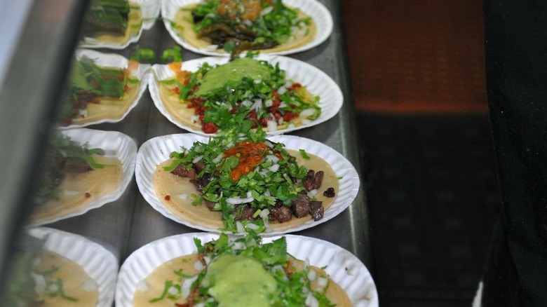 Four tacos lined up on paper plates on metal counter