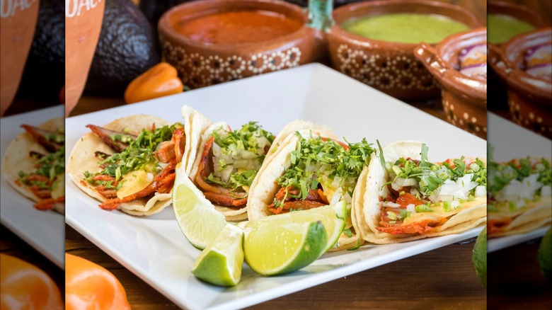 Four tacos on a white plate with toppings, lime wedges and sauces