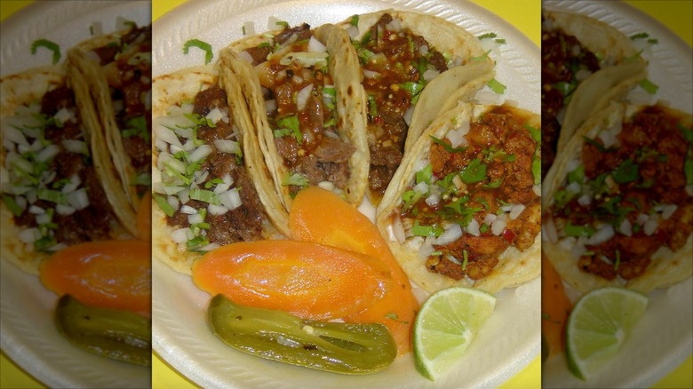 Four tacos on paper plate with toppings and pickled veggies